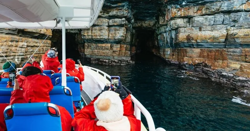 Passengers in red spray jackets on a boat inside a sea cave, seen from behind